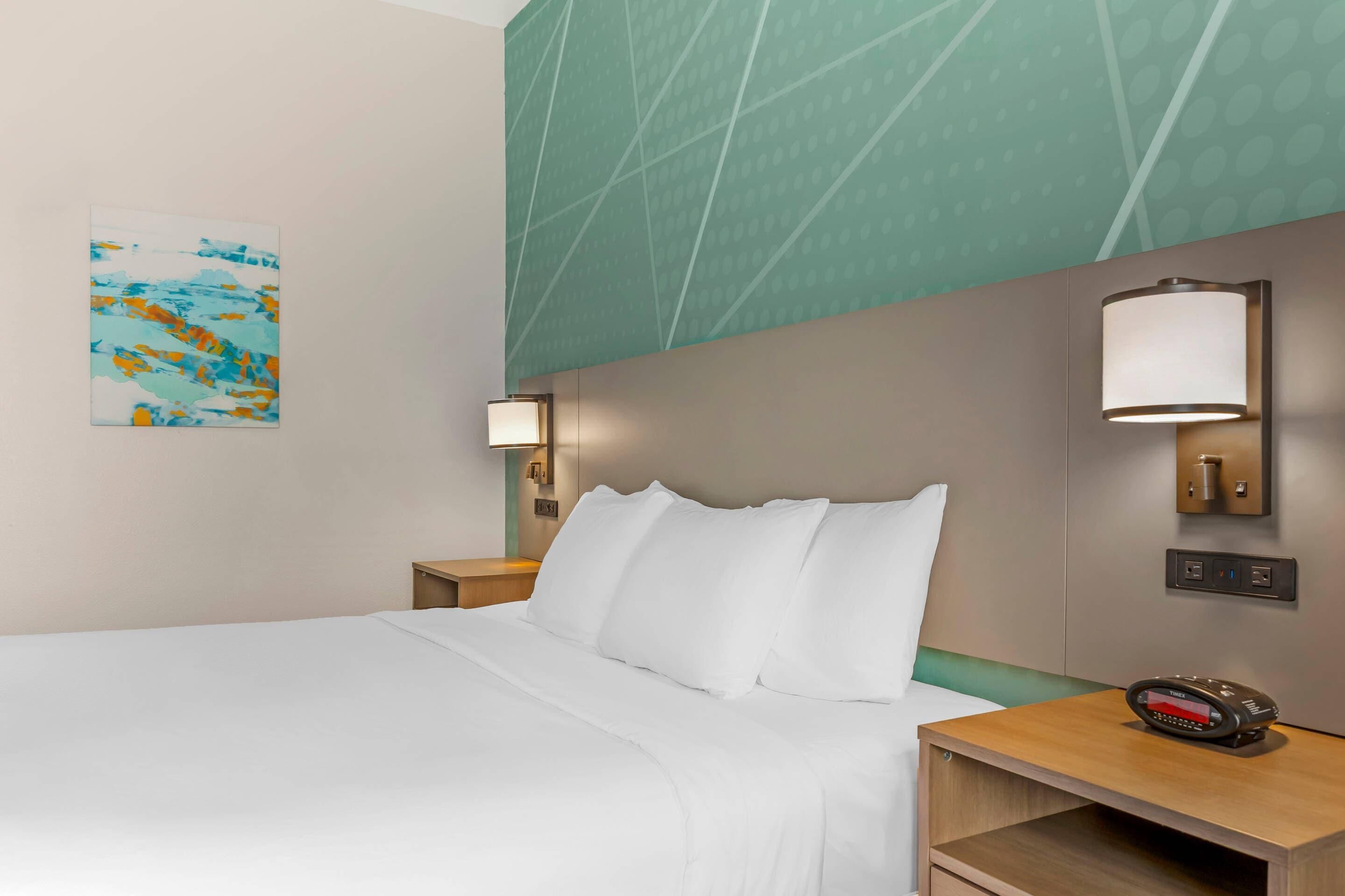 A hotel room with a large bed, a nightstand, a wall-mounted lamp, and a painting on the wall.