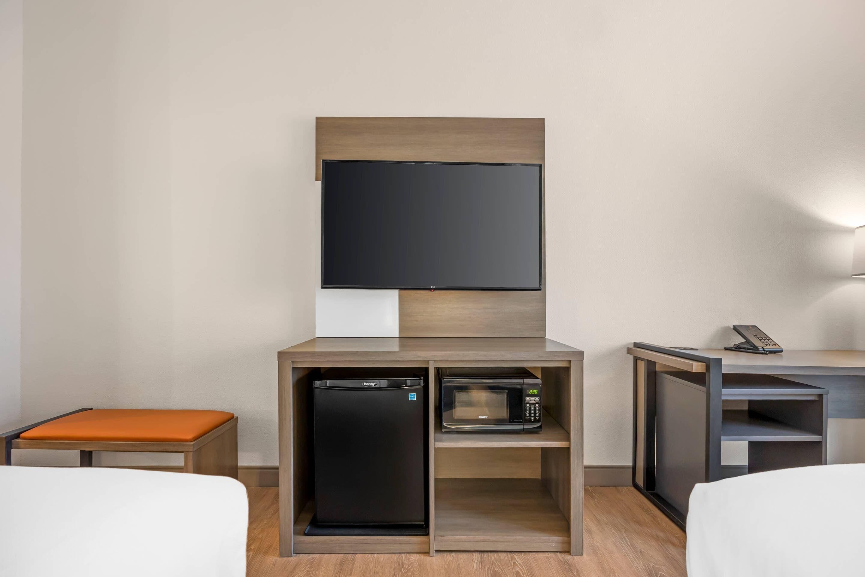 A hotel room with a bed, a small orange bench, a built-in refrigerator, a microwave, and a TV mounted on the wall.