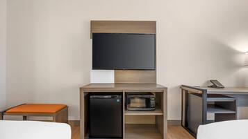 A hotel room with a bed, a small orange bench, a built-in refrigerator, a microwave, and a TV mounted on the wall.