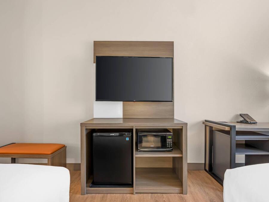 A hotel room with a bed, a small orange bench, a built-in refrigerator, a microwave, and a TV mounted on the wall.