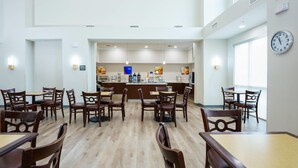 A spacious dining area with wooden tables and chairs, a central counter with a blue sign, and a clock on the wall. - Comfort Suites University Drive (College Station)