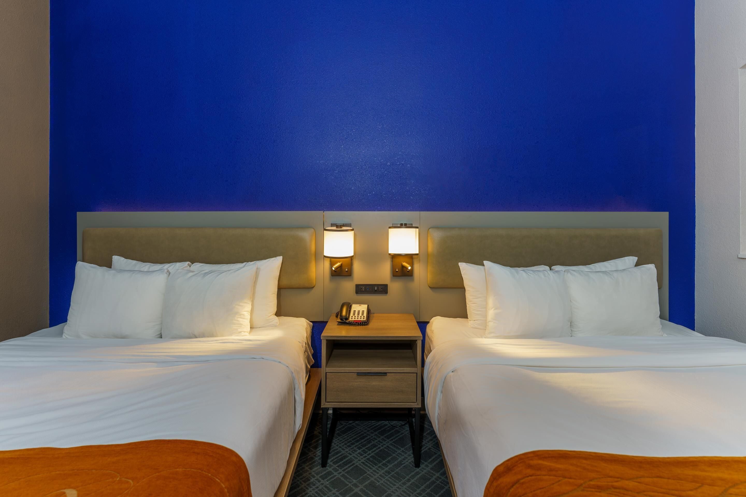 A hotel room with two beds, a nightstand with a phone, and wall-mounted lamps.