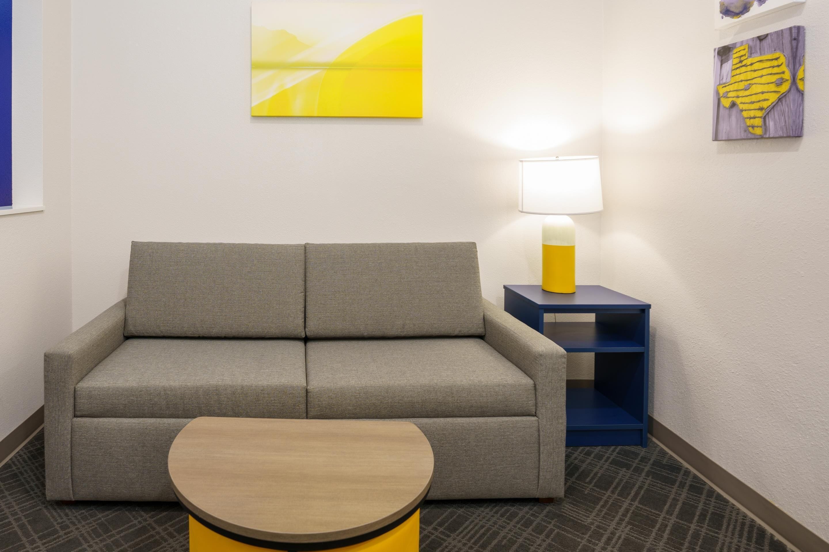Una habitación de hotel moderna con un sofá gris, una mesa de centro de madera, una lámpara sobre una mesita auxiliar azul y obras de arte en las paredes.