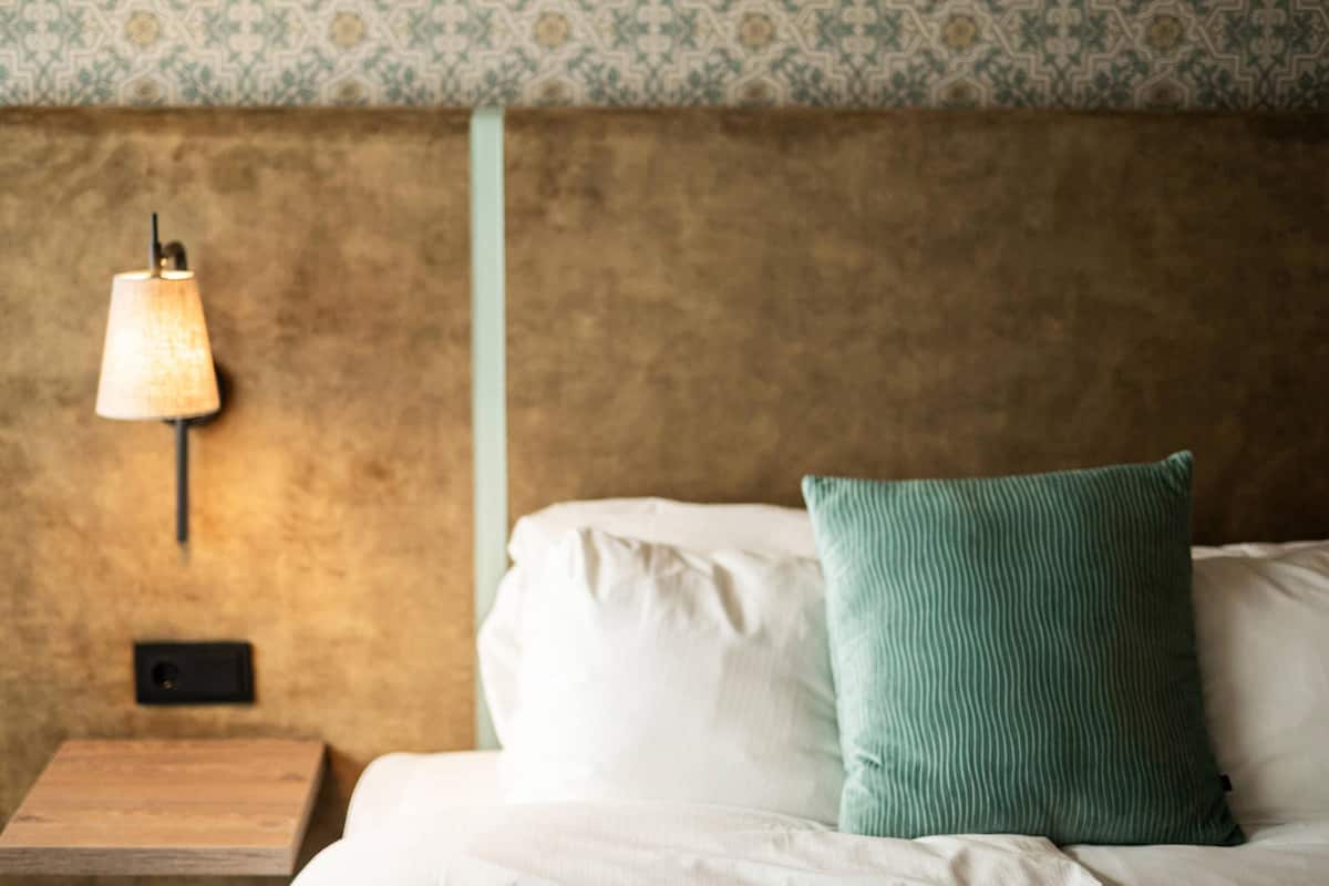 Una cama con sábanas blancas y un almohadón verde, una mesita de noche con una lámpara, y una pared con textura y papel tapiz estampado.