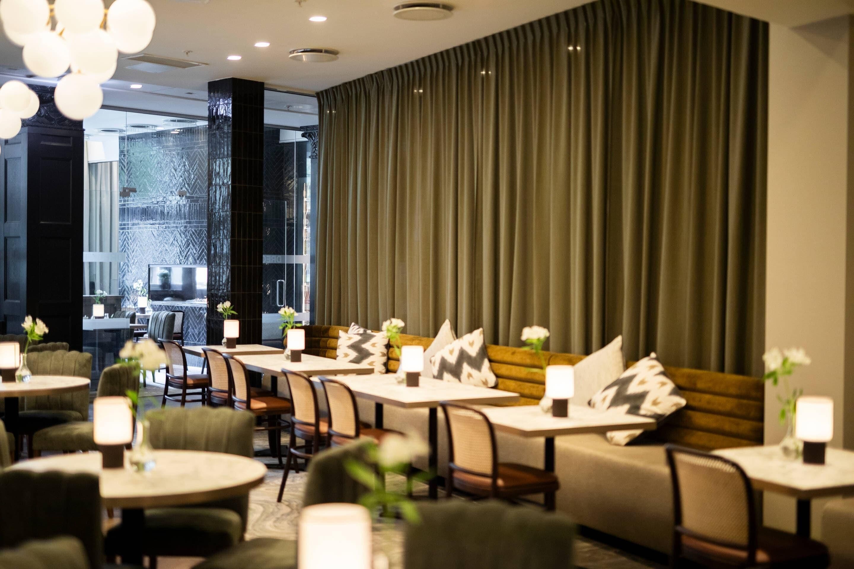 Un moderno hall de hotel con una zona de estar que incluye sofás y mesas, un ventanal con cortinas y vistas a la ciudad.