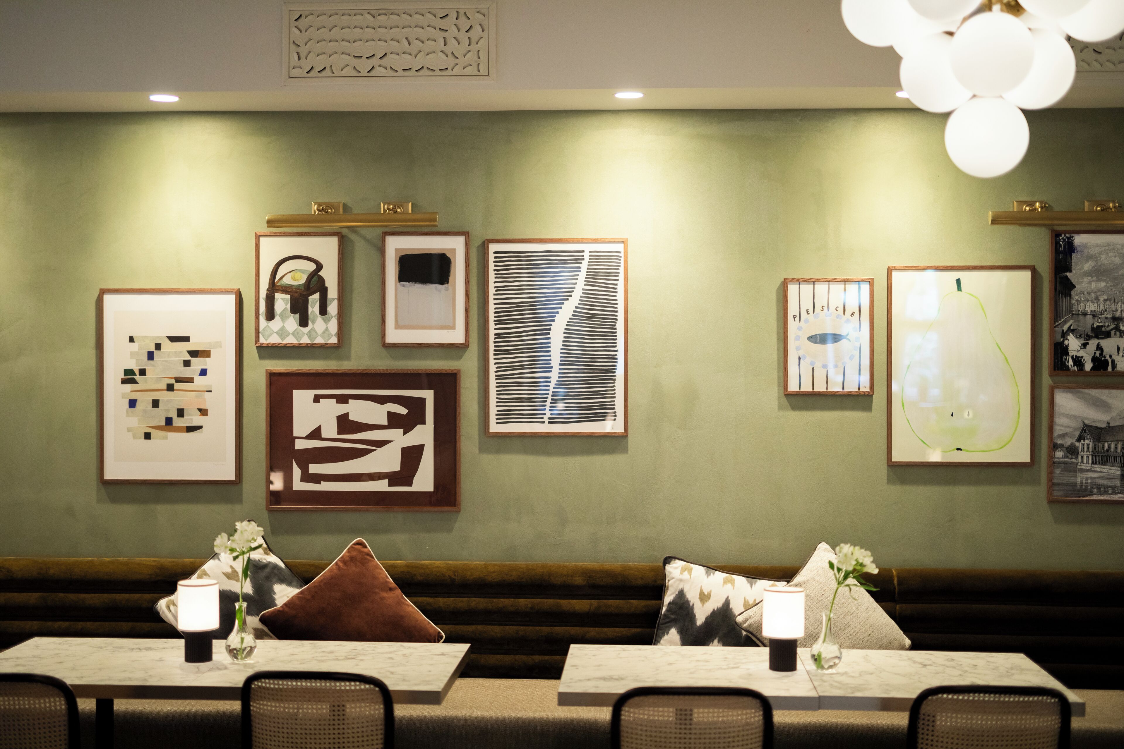 A restaurant interior with a green wall, framed artwork, a wooden bench, and tables with candles and flowers.