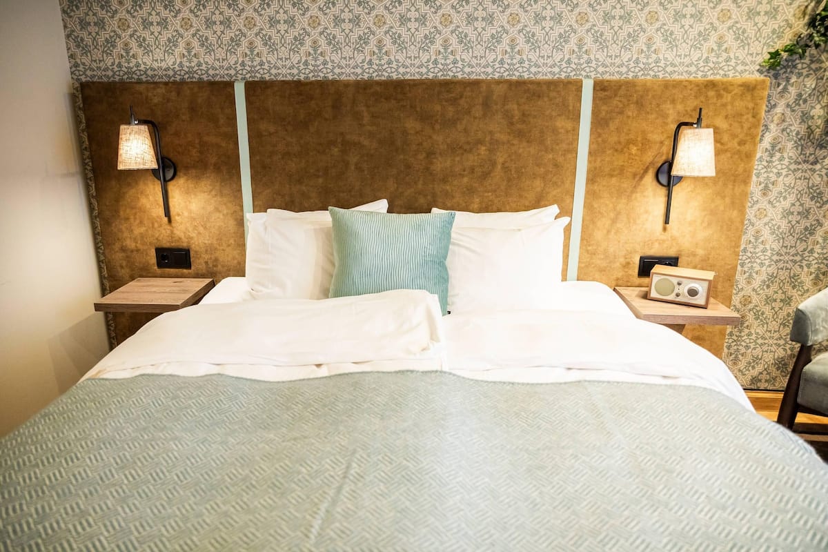 Una cama bien tendida con sábanas blancas y un plaid azul claro, un cabecero de madera, y dos mesitas de noche con lámparas y un radio.