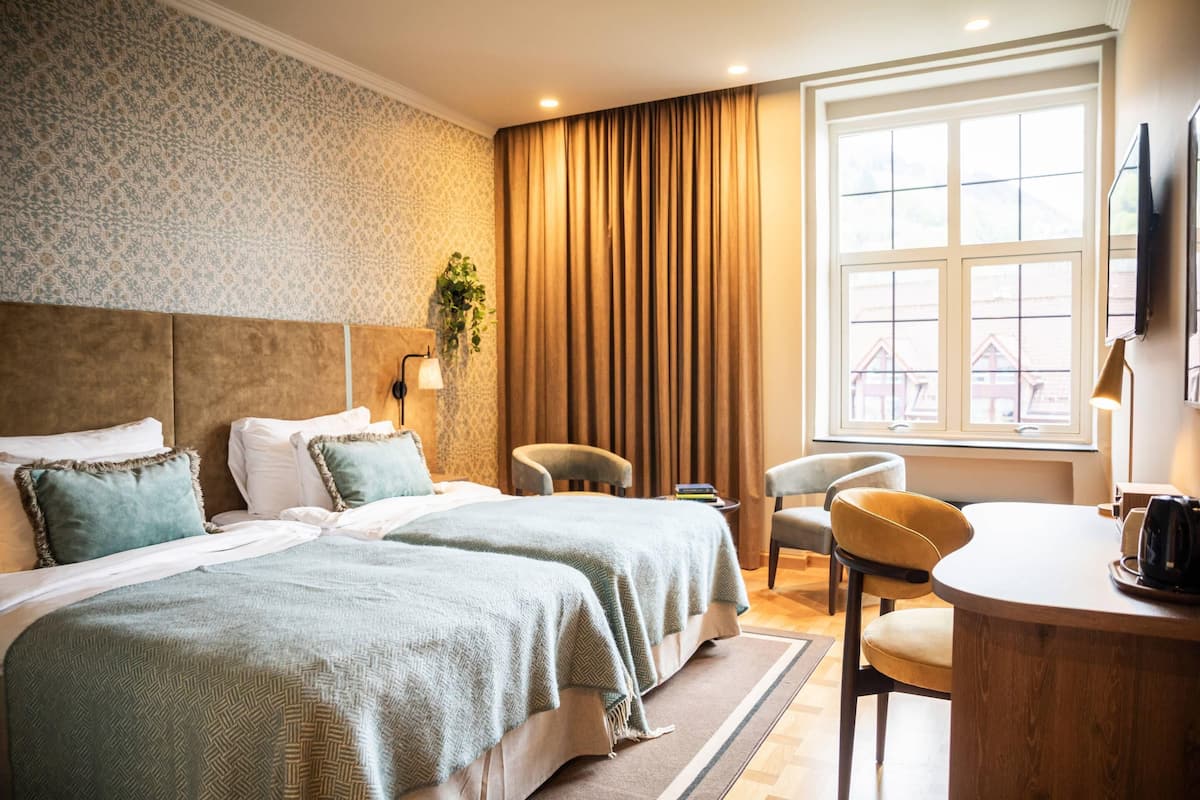 Una habitación de hotel con dos camas, un rincón de descanso con una mesa y sillas, un ventanal con cortinas y un televisor montado en la pared.