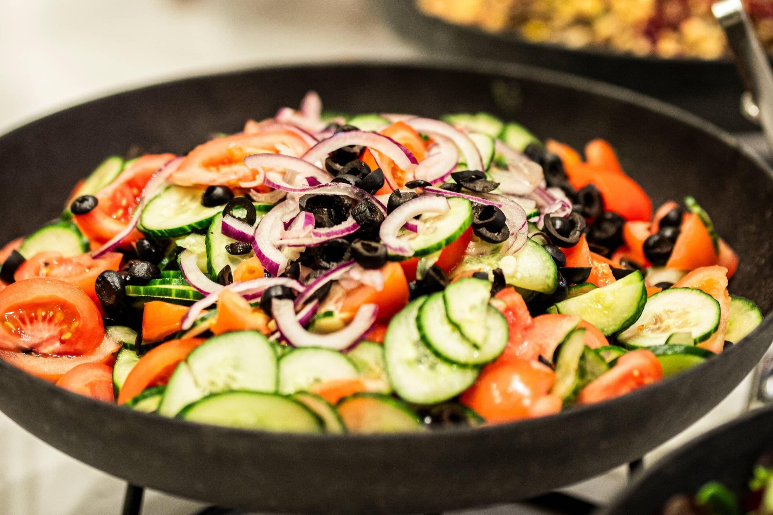 Primer plano de una ensalada de verduras colorida con tomates, pepinos, cebollas y aceitunas negras en una sartén.