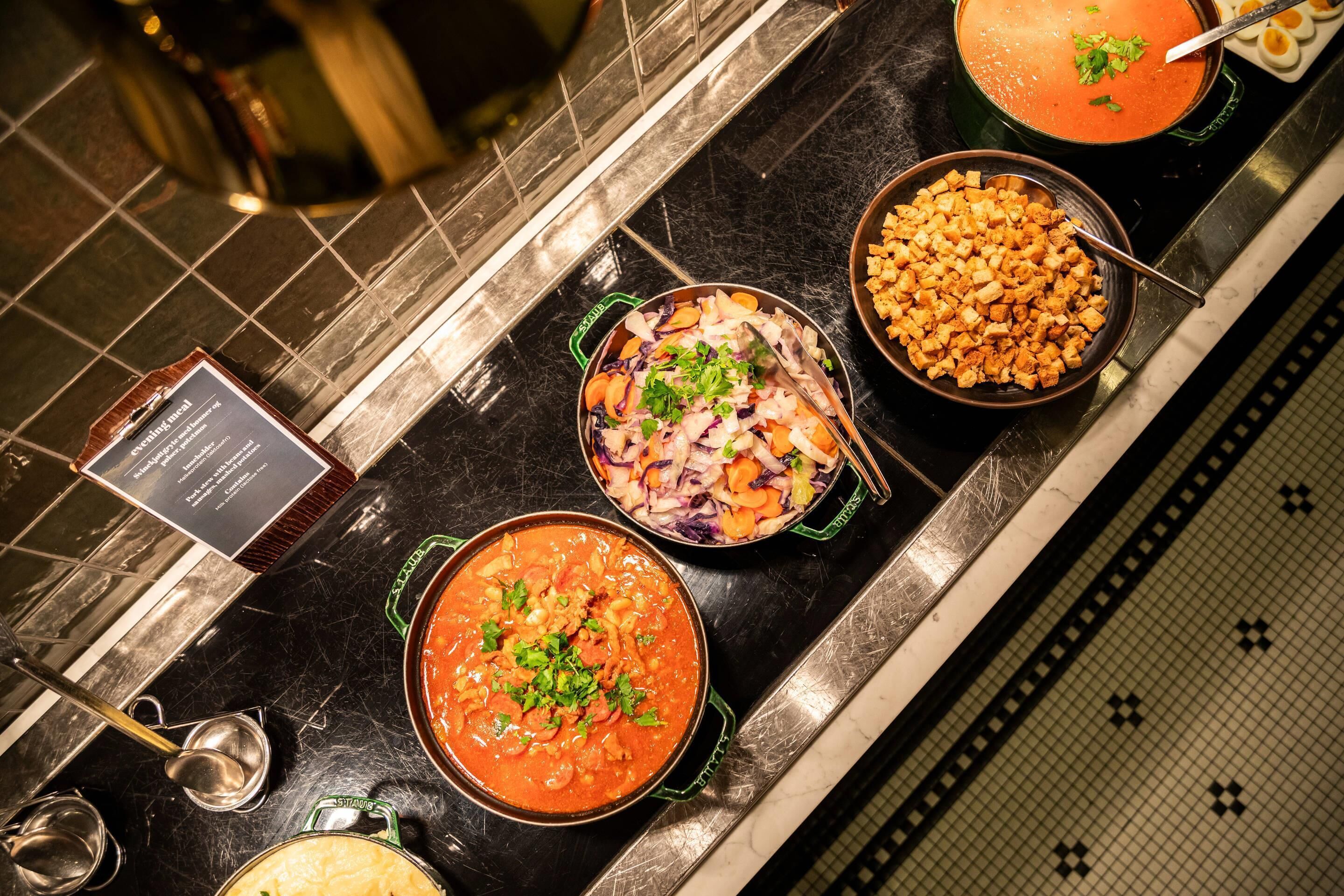 Ein Buffet mit verschiedenen Speisen, darunter eine Suppenschüssel, ein Salat und ein Gericht mit gewürfelten Gemüsesorten.