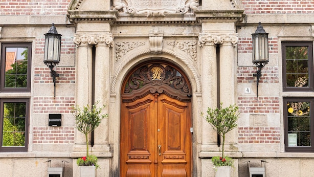 Un edificio storico con un'ampia porta in legno a due battenti, due piante in vaso e una strada acciottolata.