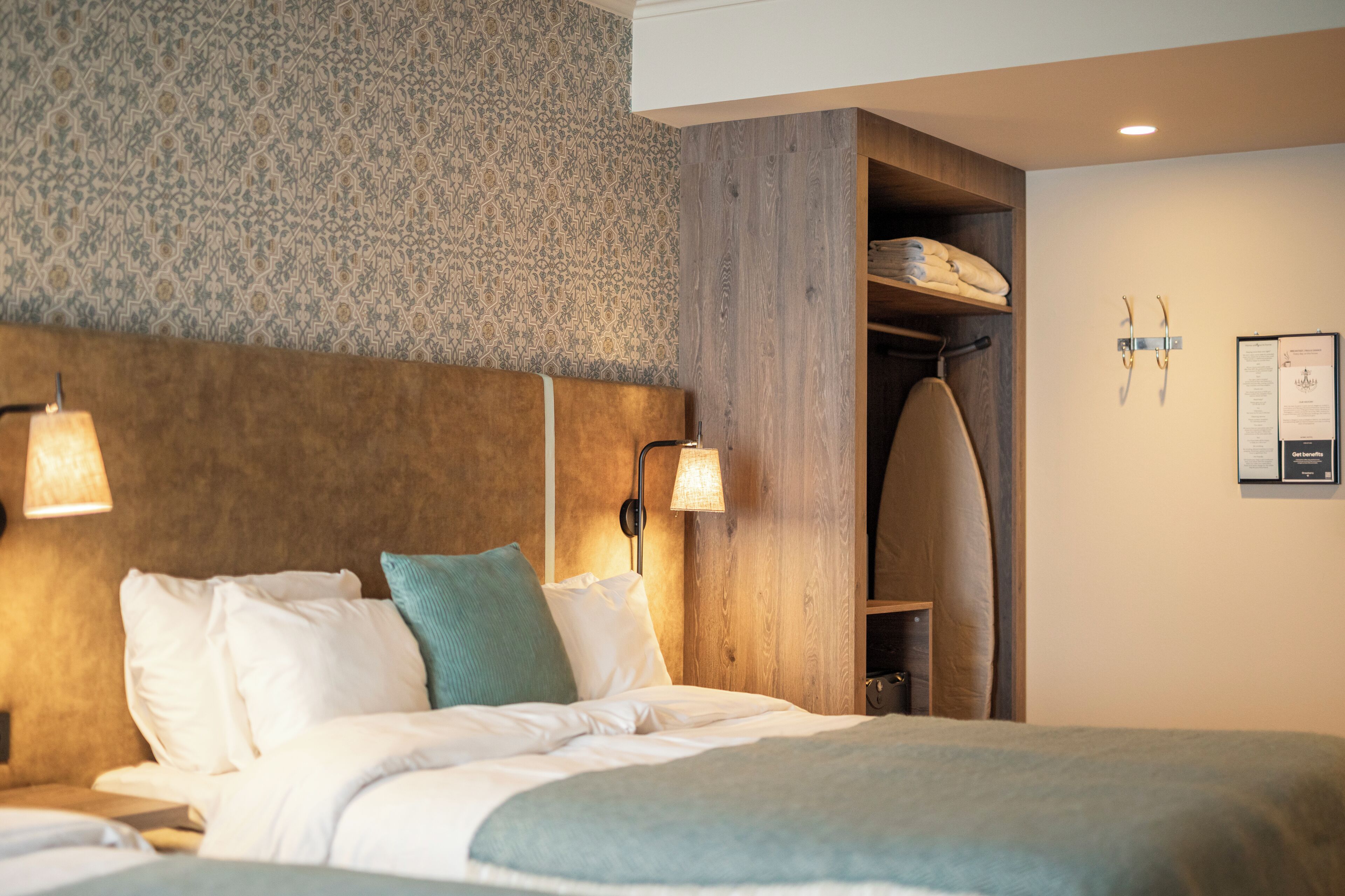 Une chambre d’hôtel comprenant un lit, deux lampes fixées au mur, une tête de lit en bois, une armoire en bois et un panneau d’information fixé au mur.