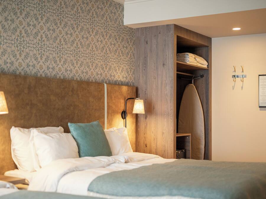 Une chambre d’hôtel comprenant un lit, deux lampes fixées au mur, une tête de lit en bois, une armoire en bois et un panneau d’information fixé au mur.