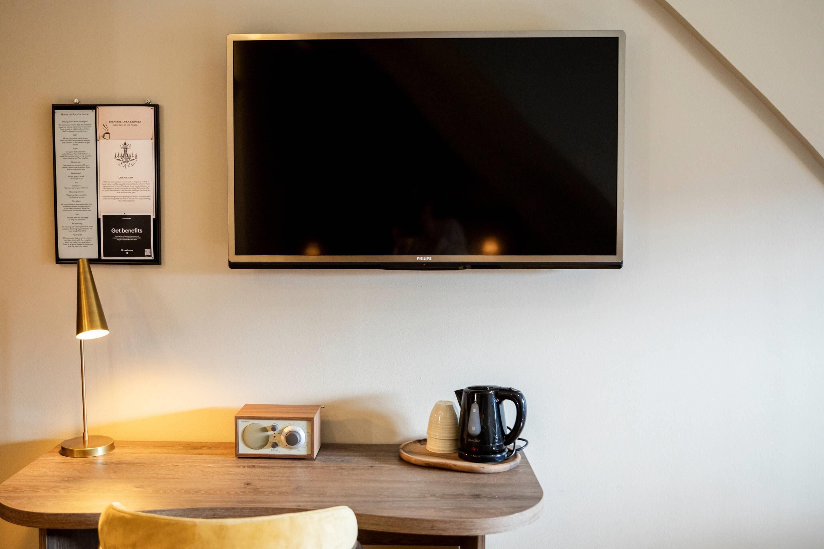 A modern room with a flat-screen TV mounted on the wall, a wooden table with a radio, a kettle, and a cup.