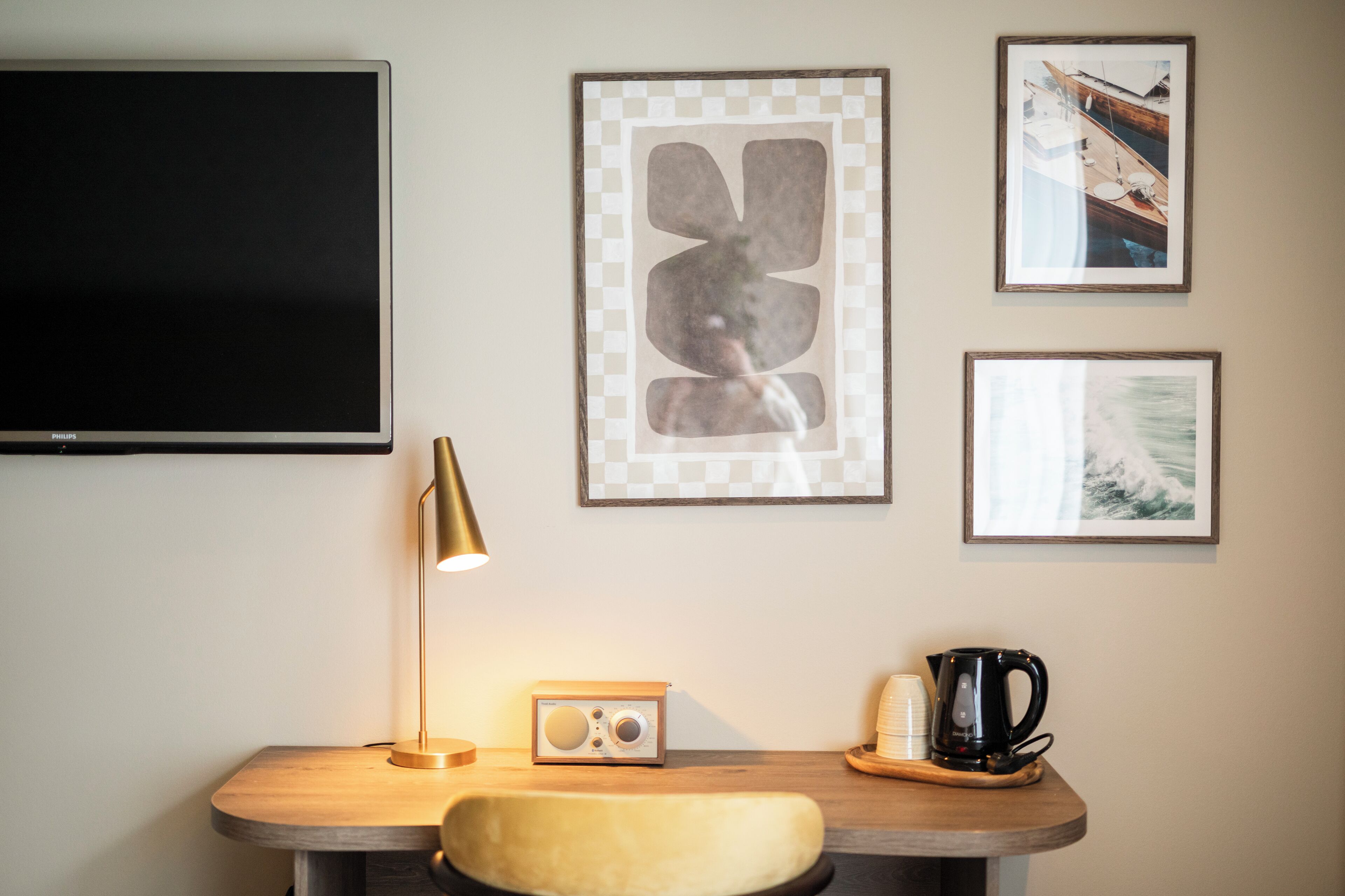 Habitación de hotel con televisor de pantalla plana, escritorio con lámpara, radio, cafetera y cuadros en la pared.