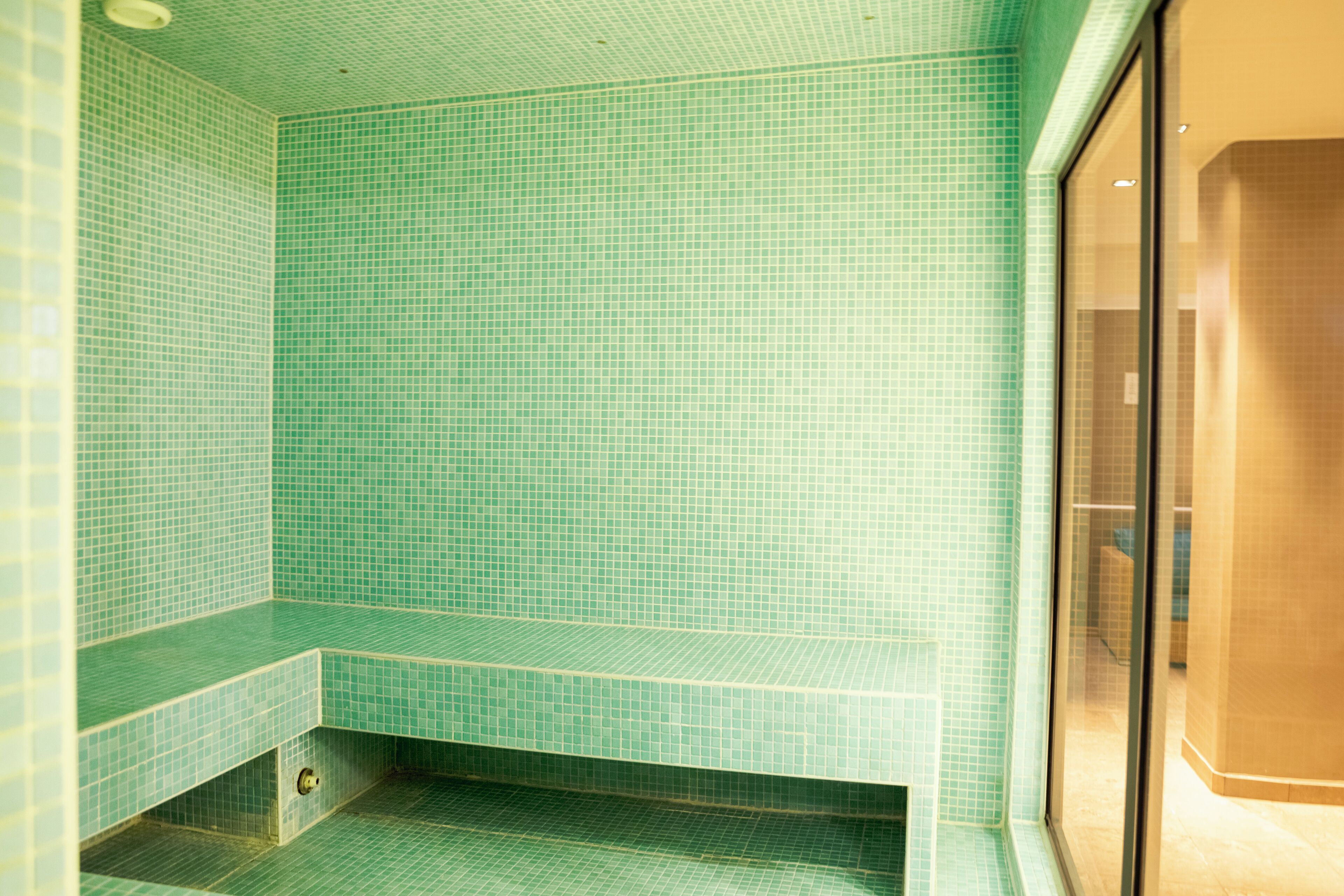 A bathroom with green tiled walls and floor, a bench, and a glass shower enclosure.