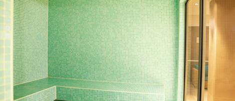 A bathroom with green tiled walls and floor, a bench, and a glass shower enclosure.