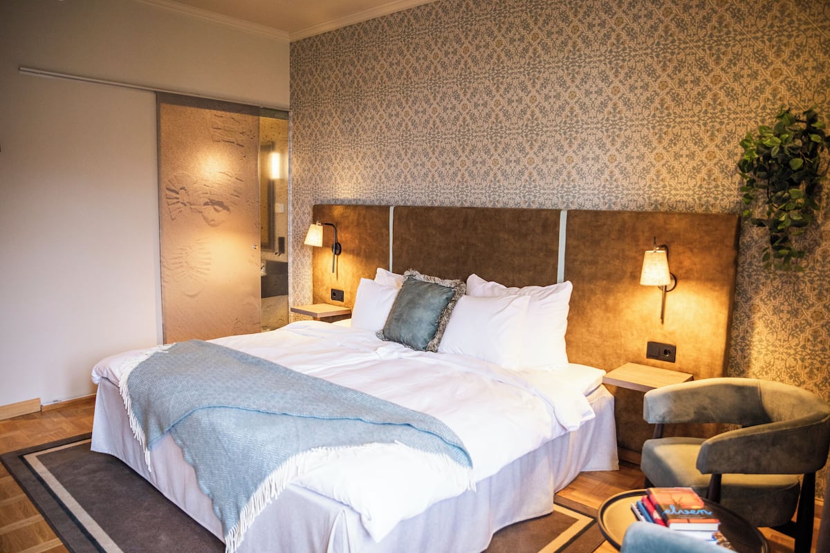 Una habitación de hotel moderna con una cama grande, dos lámparas de noche, una silla y una mesita con libros.