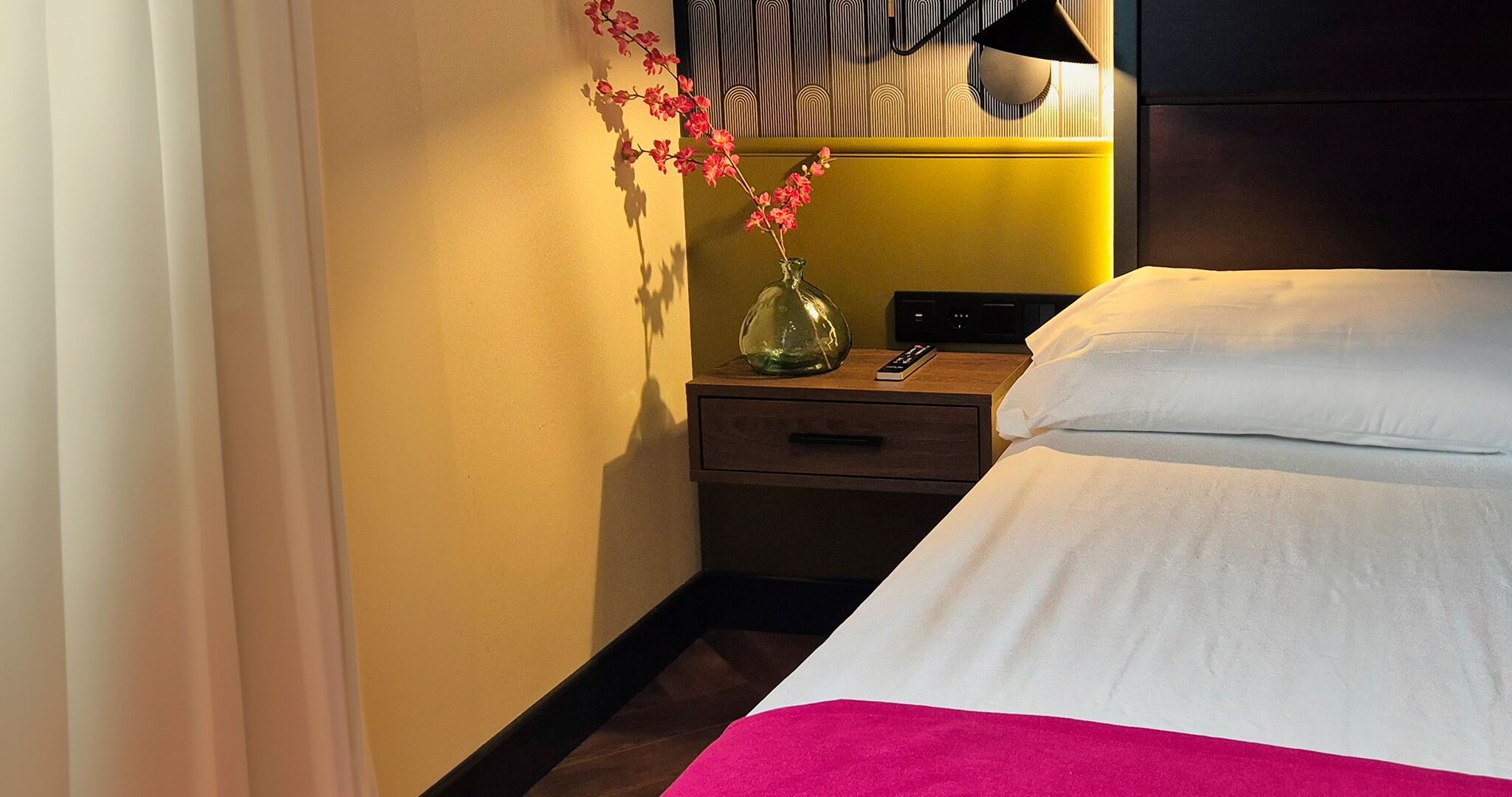 A hotel room with a bed, a nightstand, a vase with flowers, and a lamp.