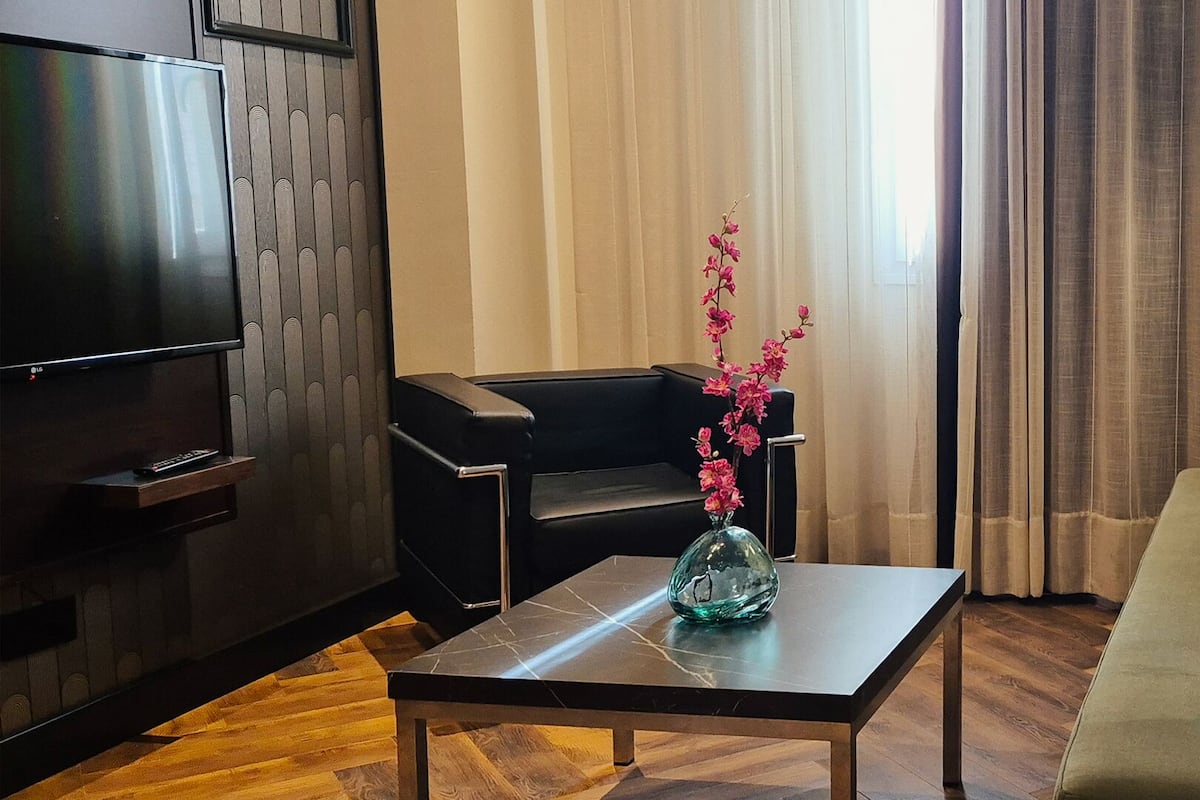 Una sala de estar moderna con un televisor de pantalla plana, un florero de cristal con flores, un sofá de cuero negro y una mesa de centro de madera.