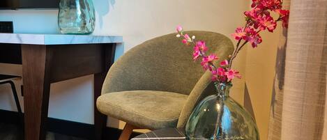 Una habitación con una butaca tapizada de verde, una mesa con cubierta de mármol y un florero de vidrio con flores rosadas.