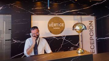 Una reception di hotel con una parete in marmo, un cartello con la scritta "VERTICE hotels" e un banco della reception.