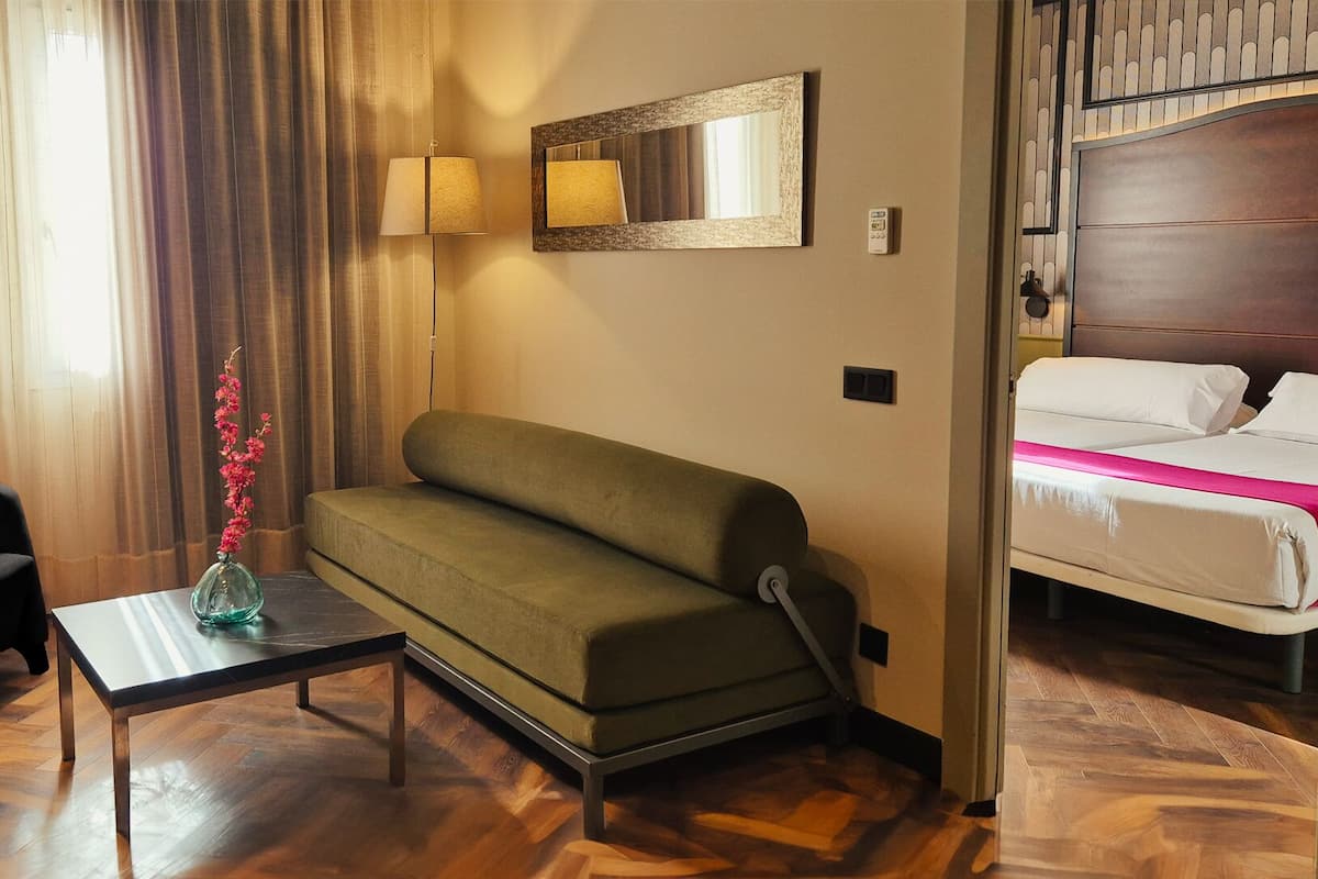 Habitación de hotel con un sofá verde, un sillón negro, una mesita con un florero y una cama con ropa blanca y cabecera de madera.