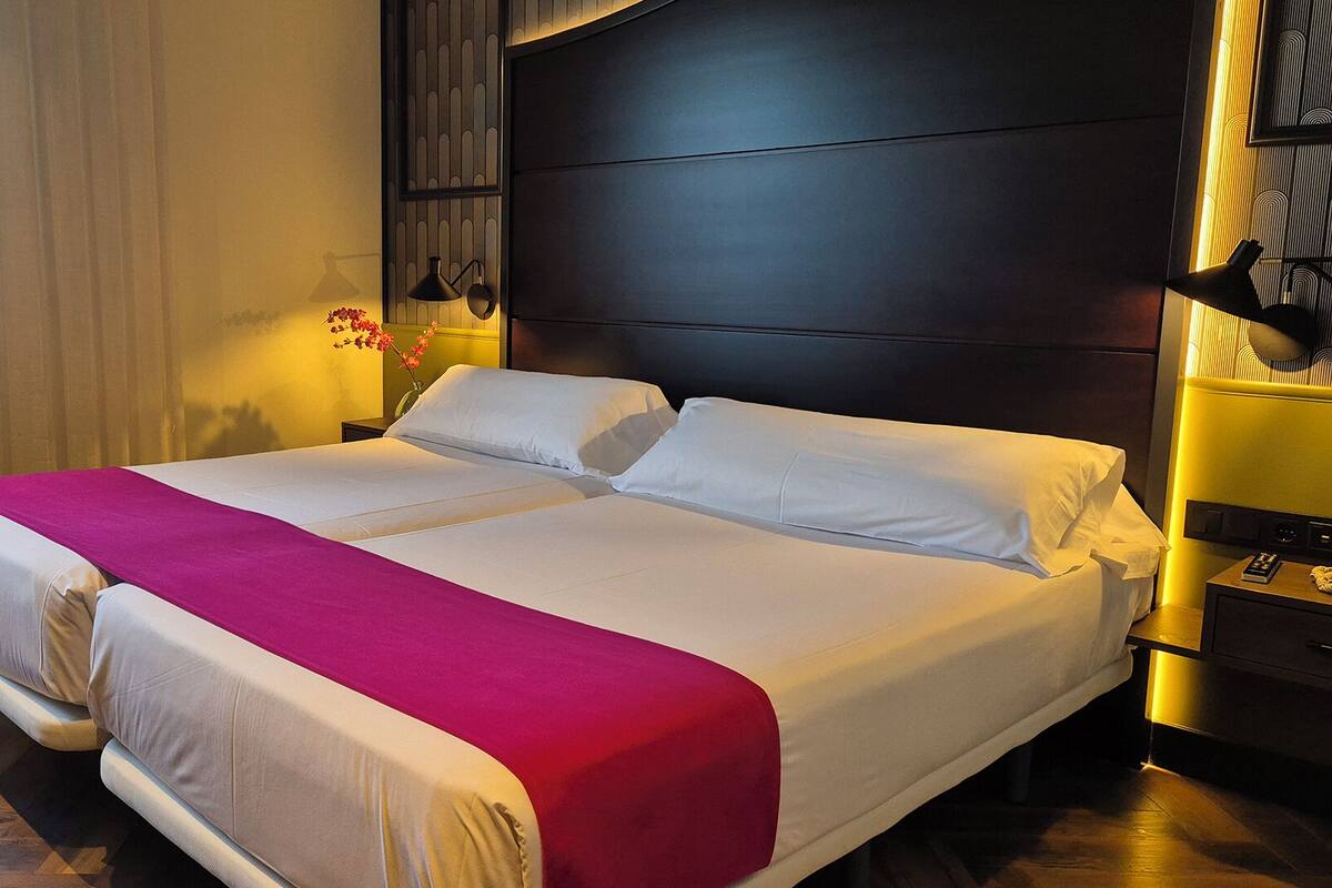 Una cama grande con ropa de cama blanca y rosa, un cabecero oscuro y una mesita de noche con una lámpara.