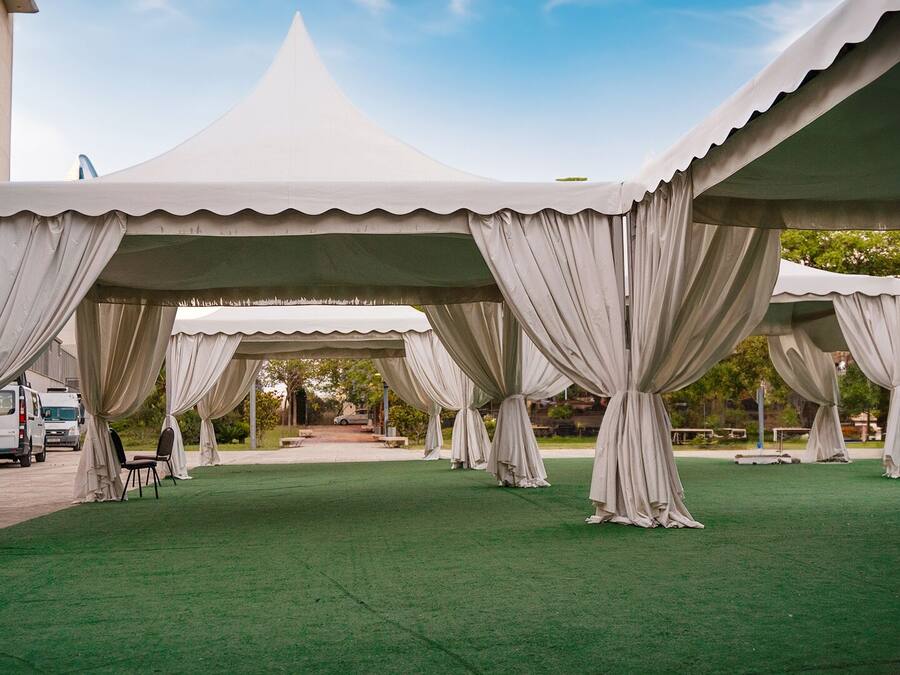 A series of white tents with draped curtains, set on a green artificial turf, with a building and parked vehicles in the background.