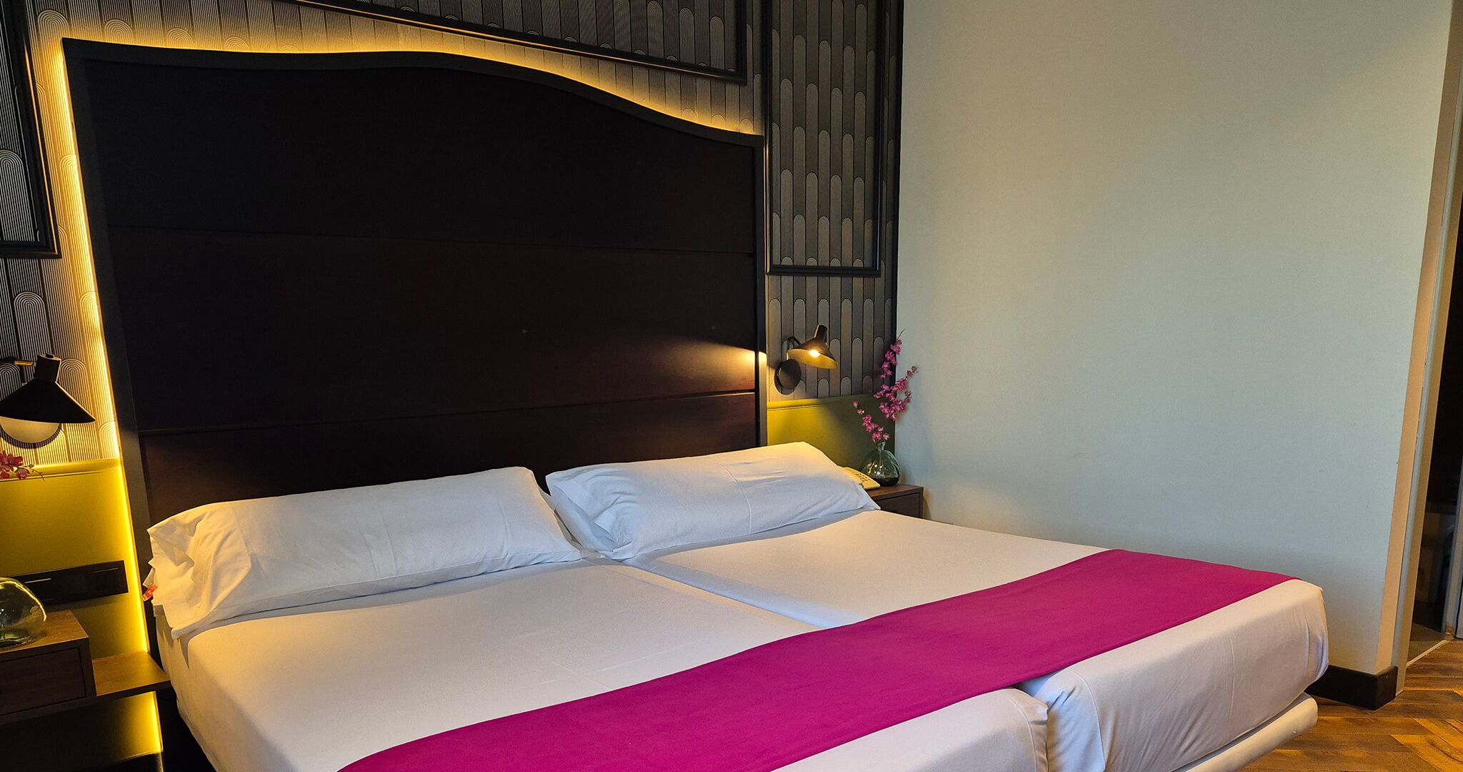 Un lit double avec du linge de lit blanc et une touche de rose, une tête de lit sombre et une lampe fixée au mur.