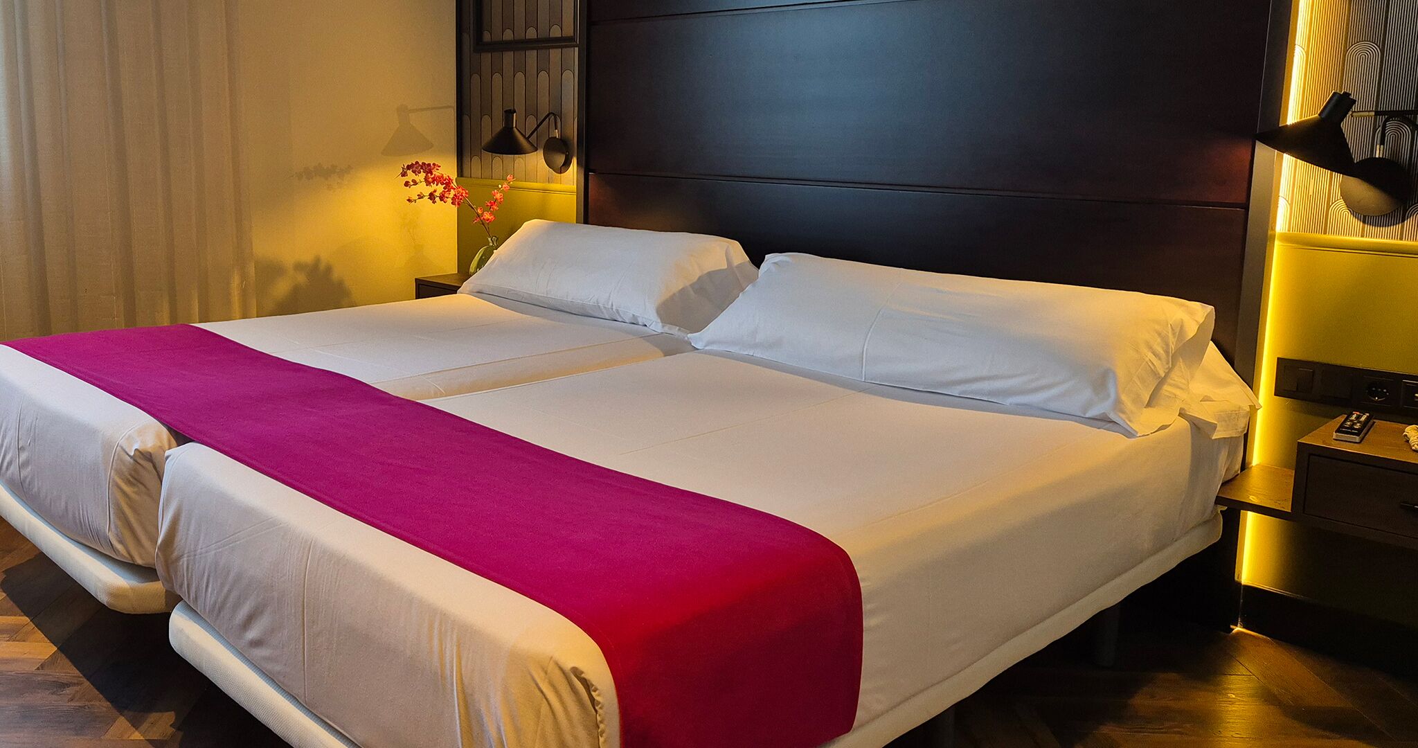 A double bed with white and red bedding, a bedside table with a lamp, and a wall-mounted light fixture.