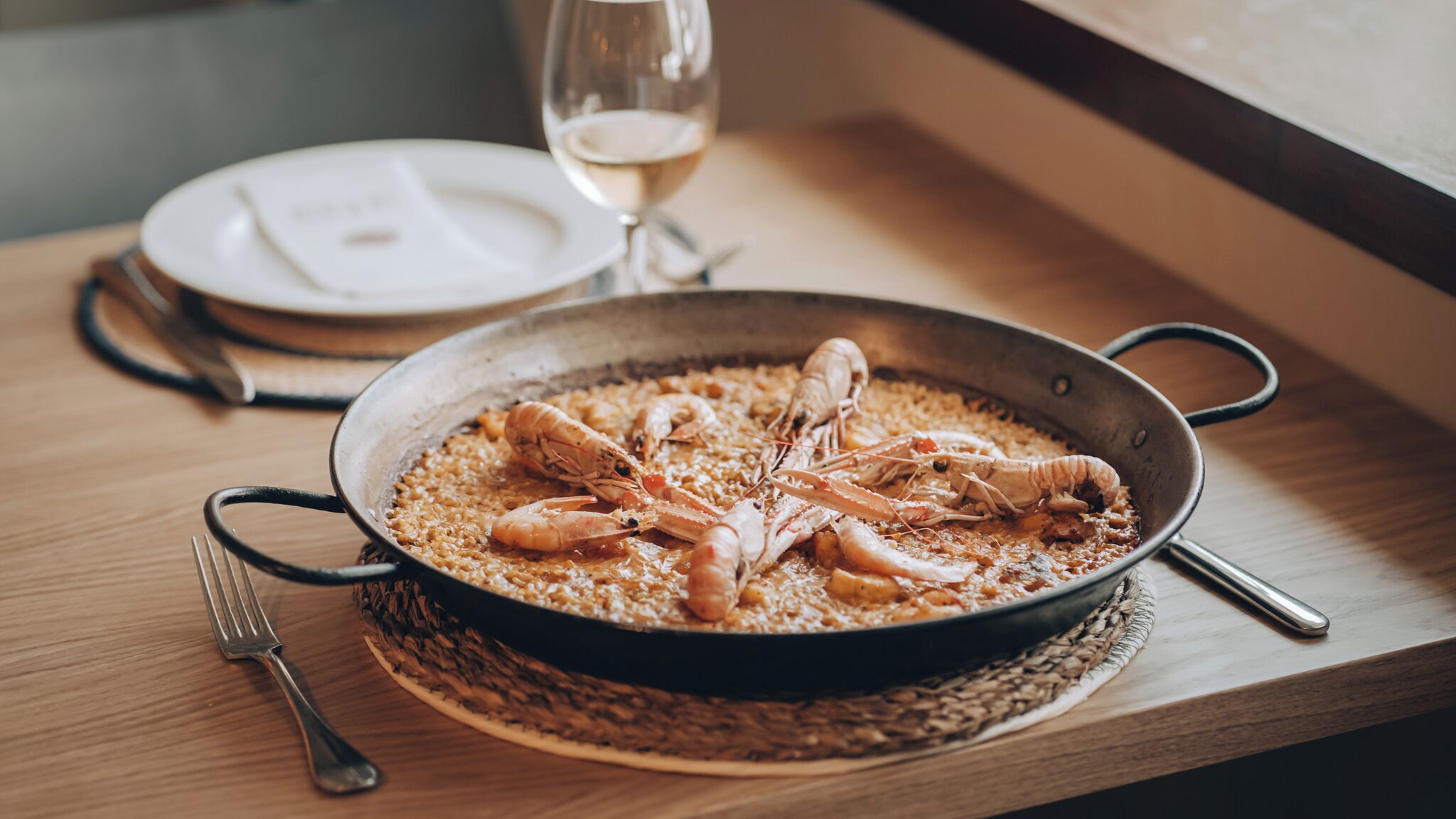 A seafood dish in a pan, accompanied by a fork and spoon, on a wooden table with a glass of white wine in the background.