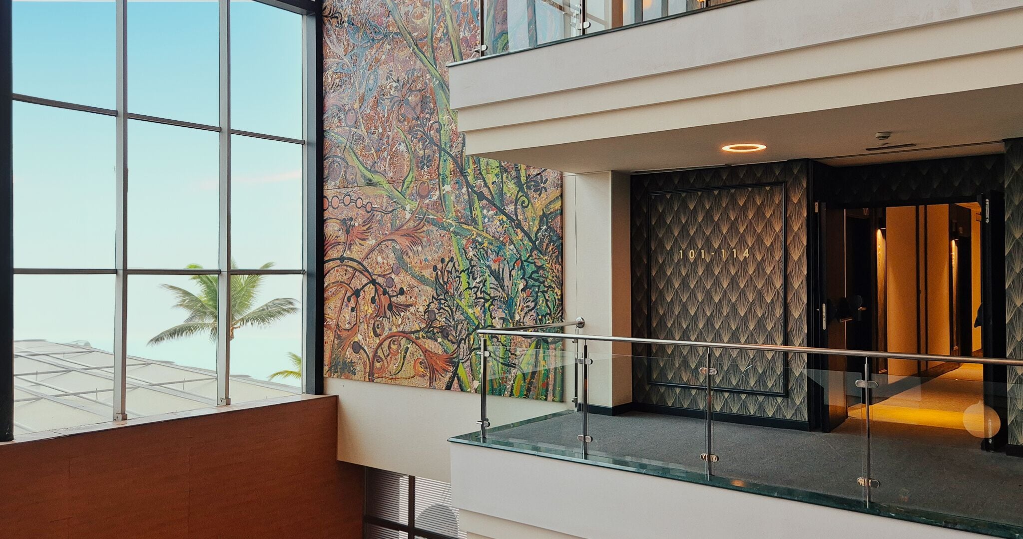 Un moderno vestíbulo de hotel con ventanas amplias, un mural abstracto de colores y una barandilla de vidrio.