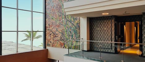 Un moderno vestíbulo de hotel con ventanas amplias, un mural abstracto de colores y una barandilla de vidrio.