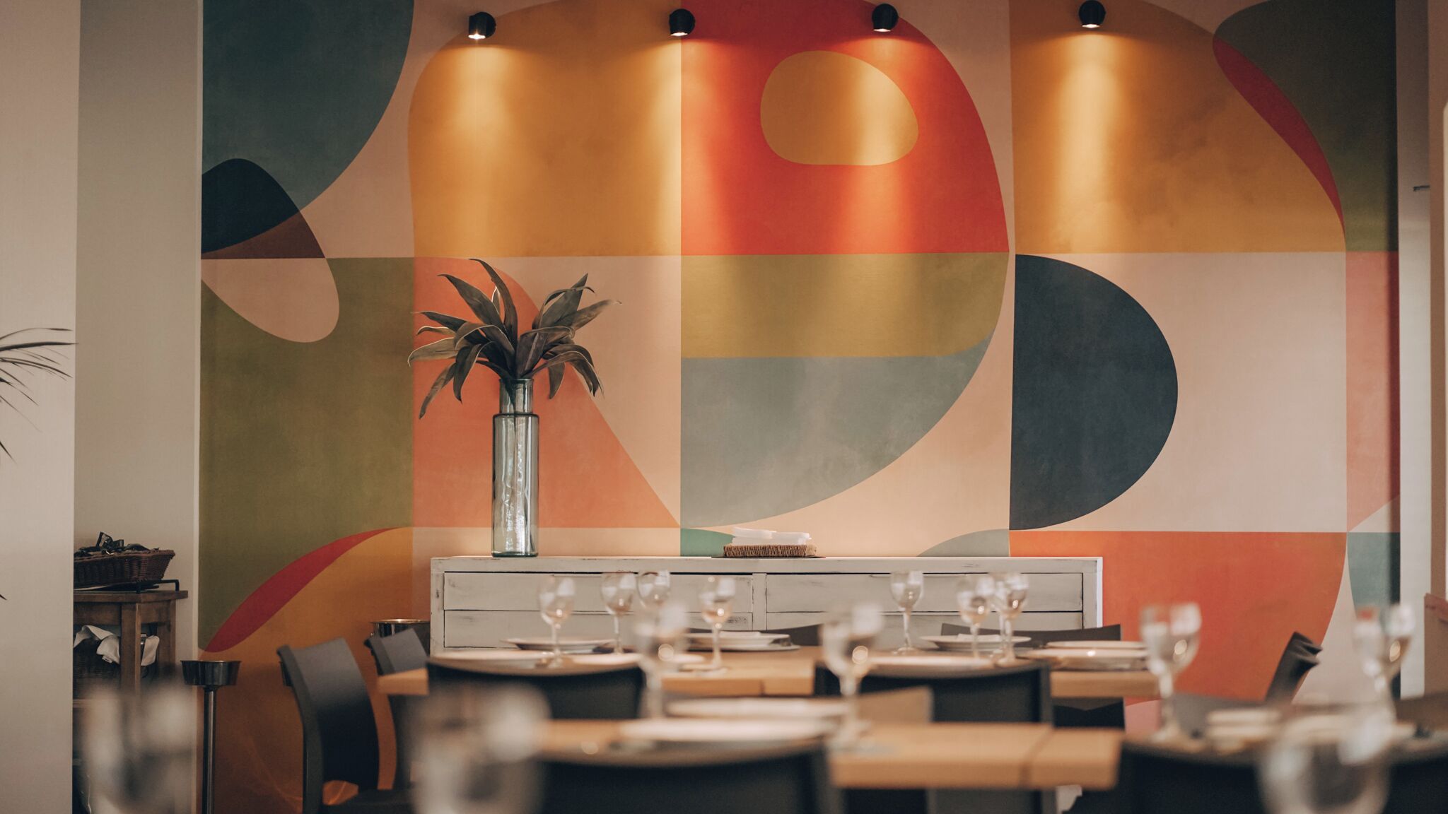 A dining area with a modern table setup, a vase with a plant, and a colorful abstract wall design.