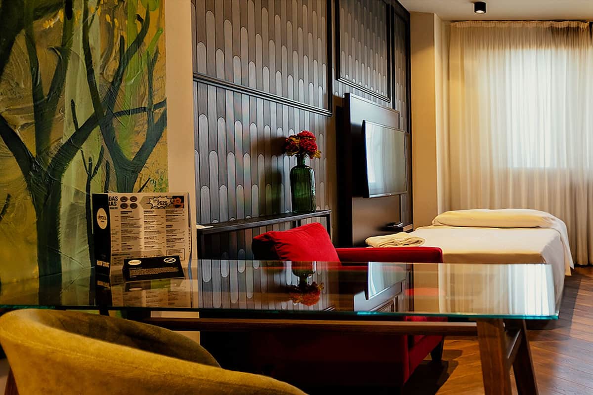 Habitación de hotel con un escritorio de vidrio, dos camas, un televisor y un ventanal con cortinas.
