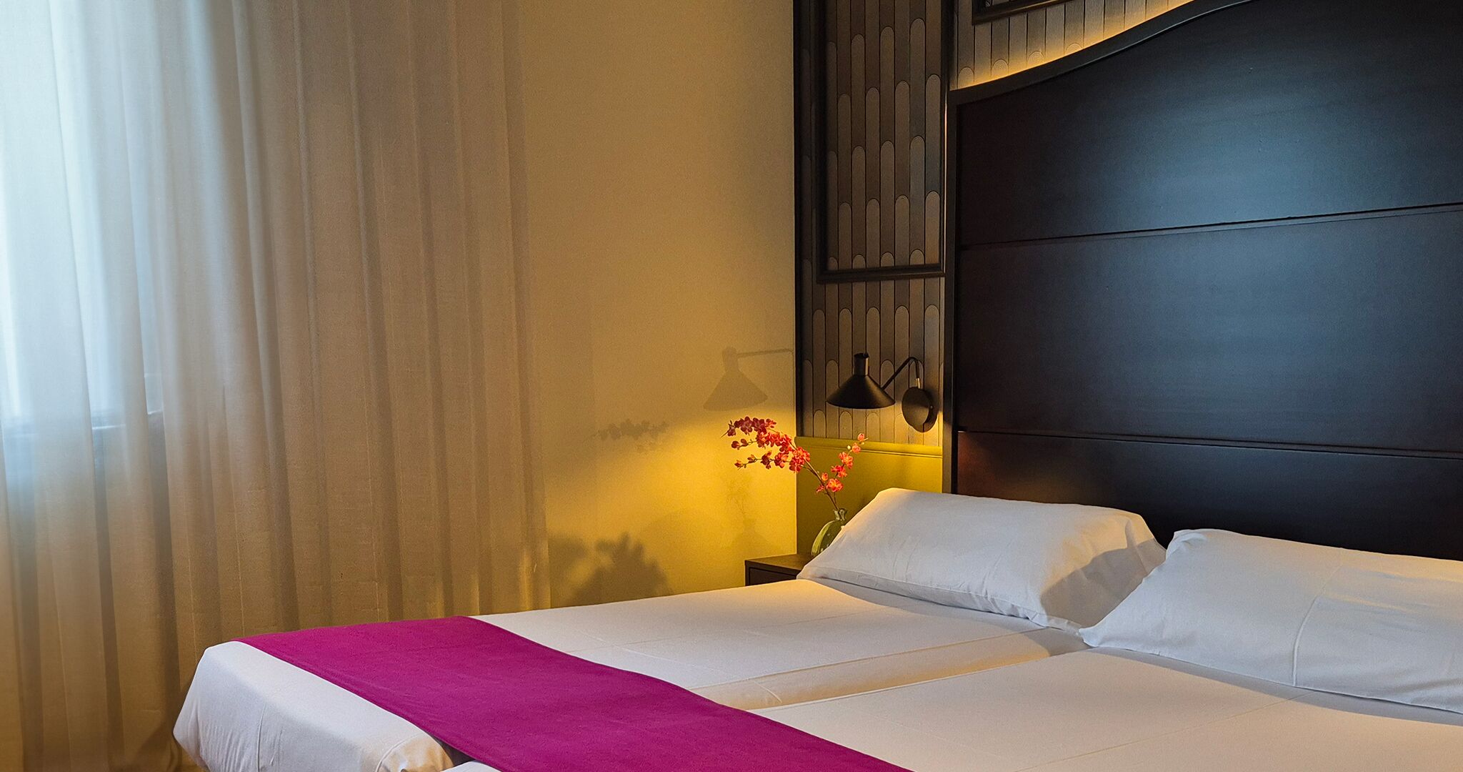 Quarto de hotel com uma cama grande, mesa de cabeceira com abajur e janela com cortinas.