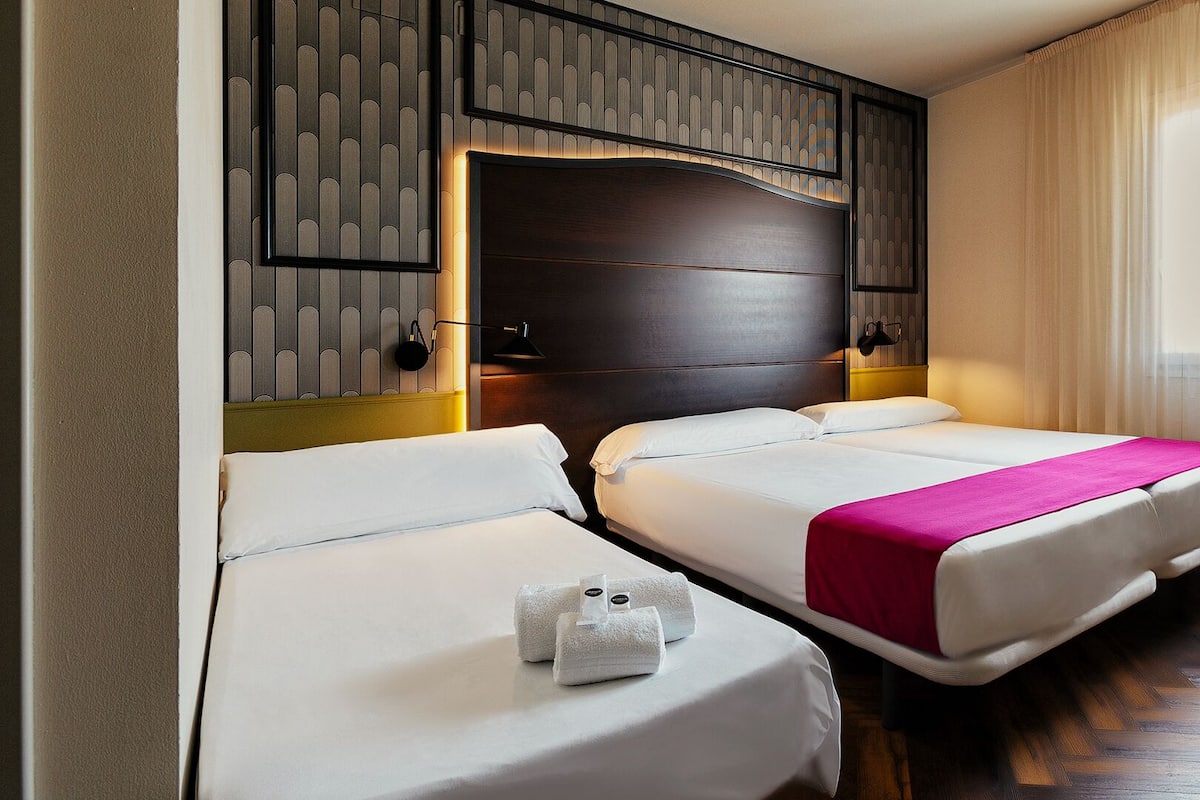 Una habitación de hotel con dos camas, cabecera de madera, un ventanal con cortinas translúcidas y suelo de madera.