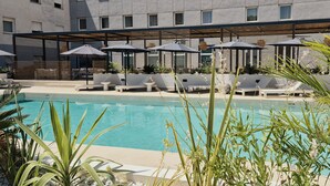 A pool area with lounge chairs and umbrellas, surrounded by plants and a building with large windows. - Hotel Vértice Aljarafe (Bormujos)