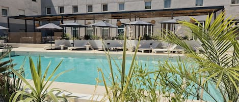 Un área de piscina con sillones de descanso y sombrillas, rodeada de plantas y un edificio con amplias ventanas.