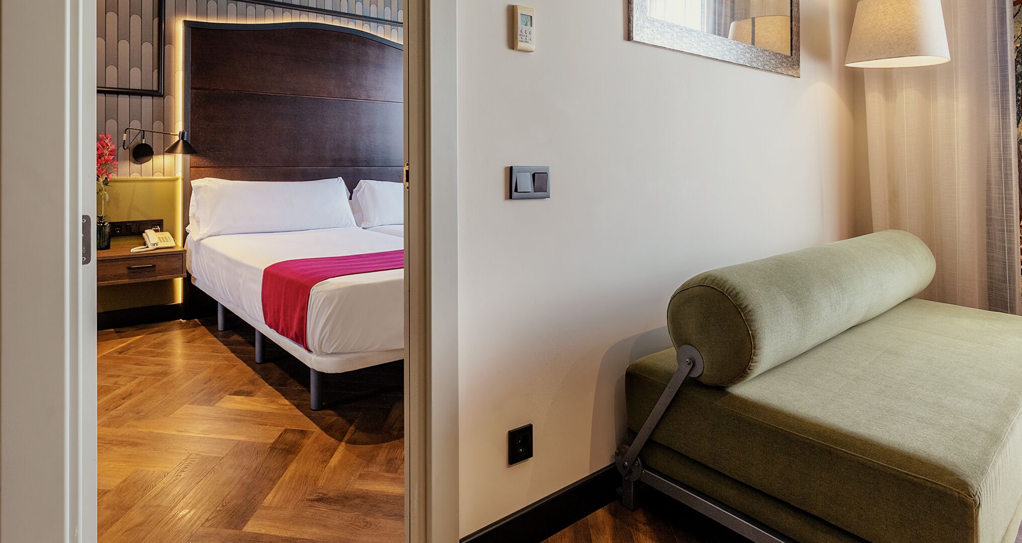Habitación de hotel con cama, mesita de noche, lámpara y sofá verde.