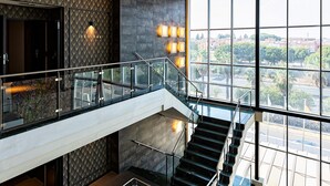 A modern interior with a glass staircase, a patterned wall, and a city view through large windows. - Hotel Vértice Aljarafe (Bormujos)