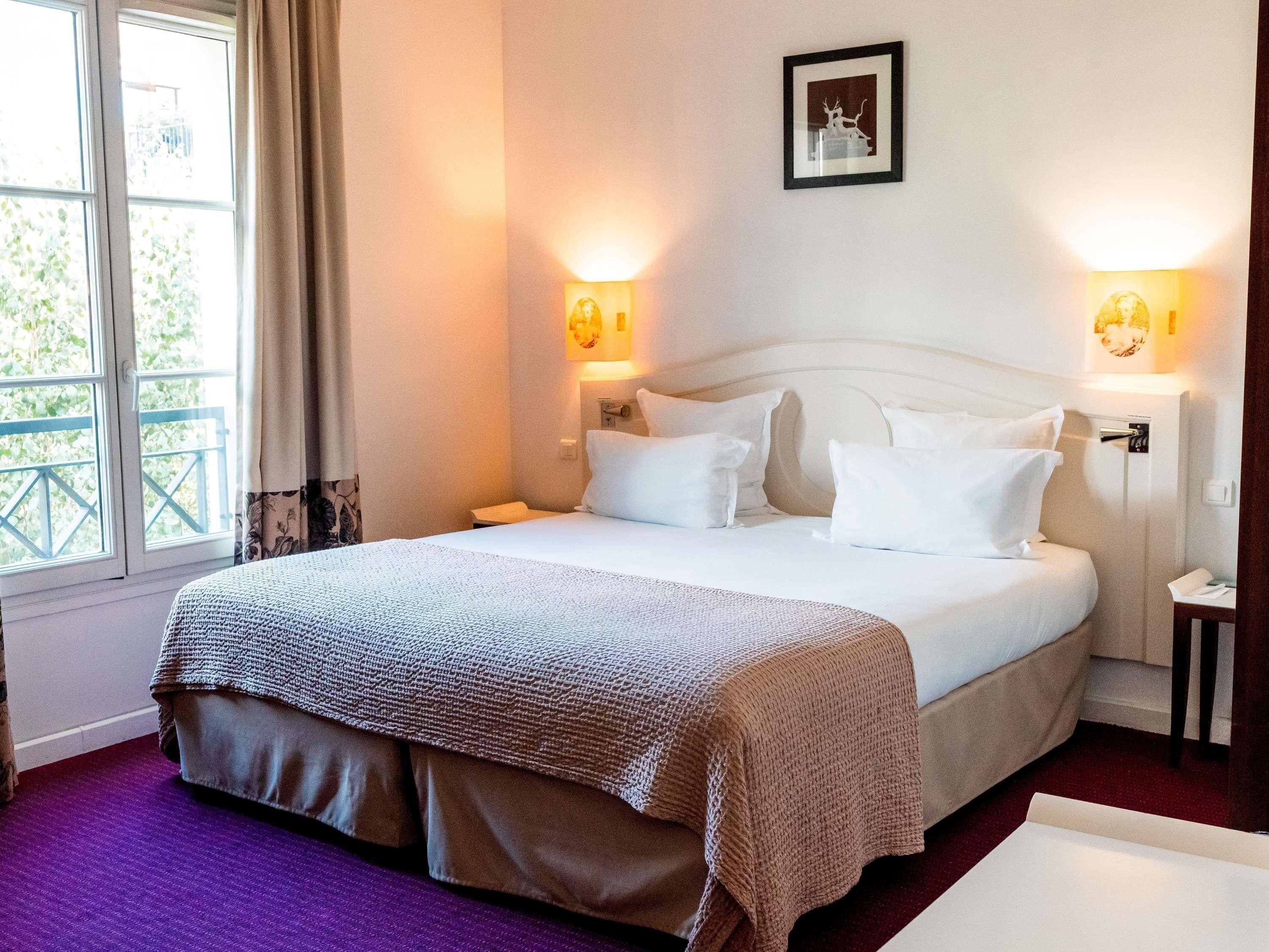 A hotel room with a bed, two bedside tables, a window with curtains, and wall-mounted lights.