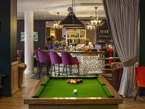 Ein Billardtisch mit grüner Spielbahn, eine Poolqueue und farbige Kugeln in einem Raum mit Barbereich, violetten Barhocker und einer Kristallleuchter.