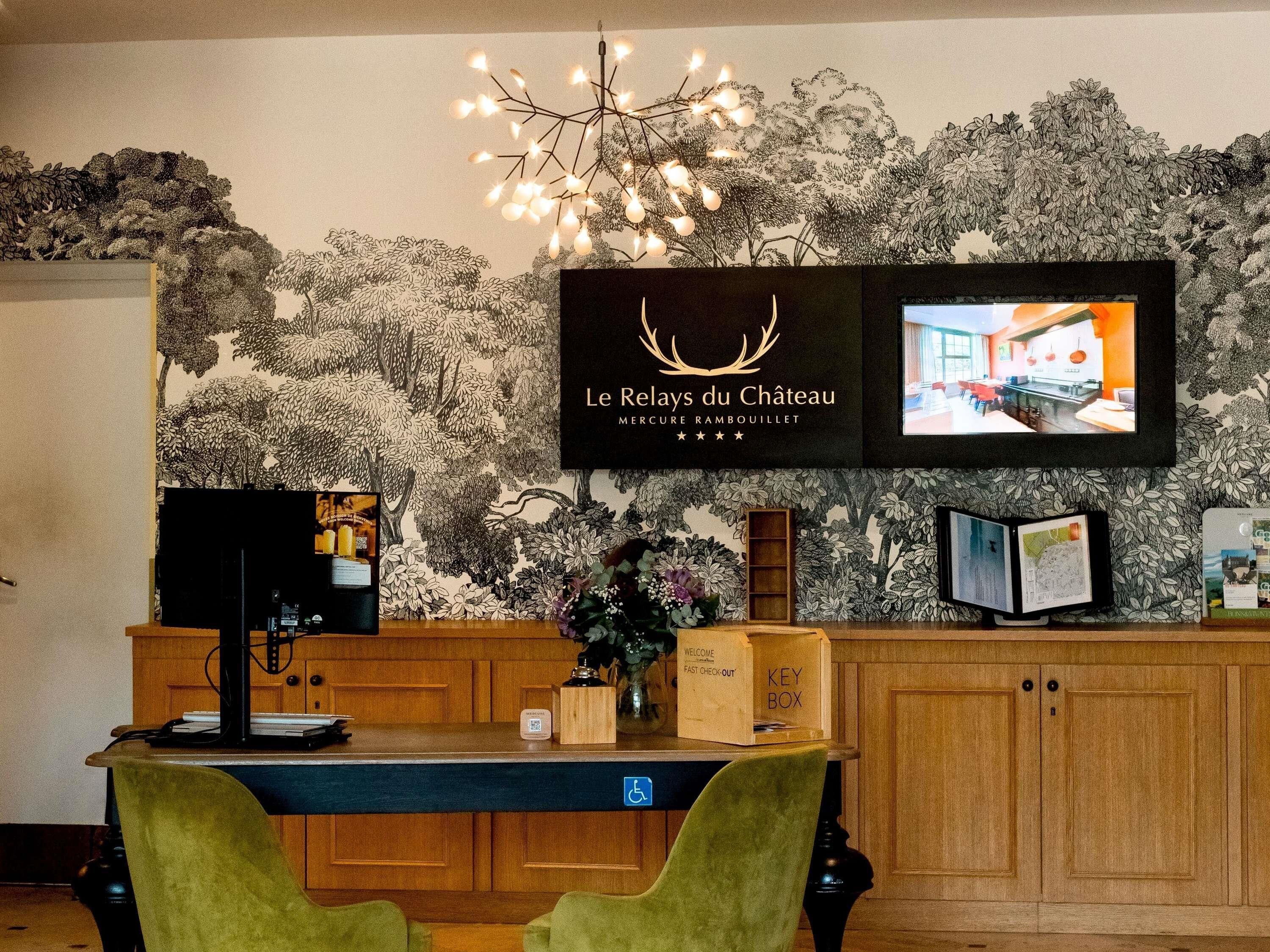 Tauleller de recepció amb un monitor que mostra l'interior d'un restaurant, una làmpada de cristall i un mural a la paret.