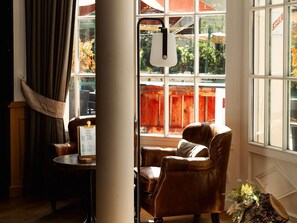 A room with a leather armchair, a table with a menu, and a window with a view of a wooden fence and greenery. - Mercure Rambouillet Relays du Chateau (Rambouillet)