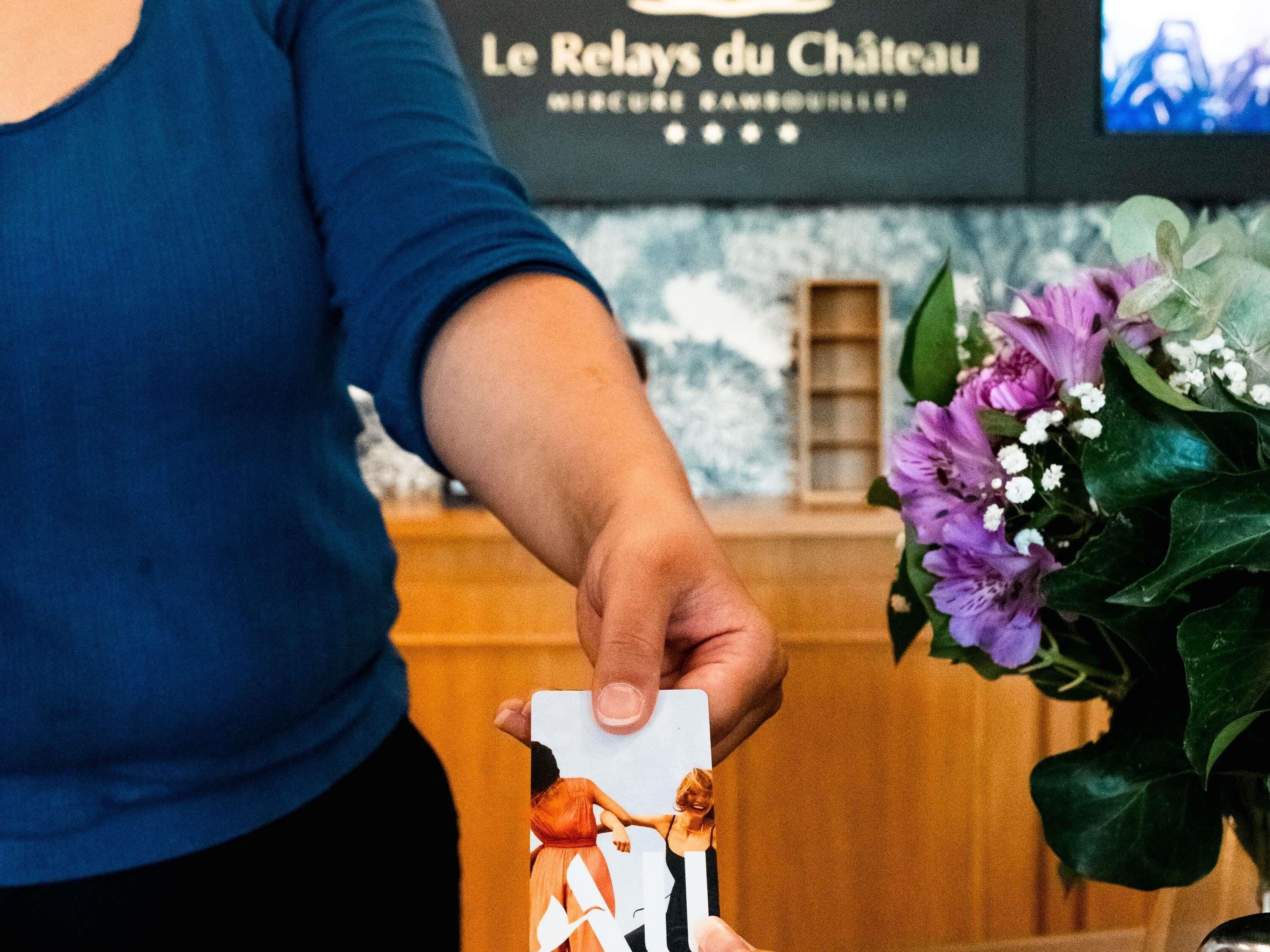 Une personne tenant une carte ornée d’un motif floral, debout devant une enseigne sur laquelle on peut lire « Le Relais du Château ».