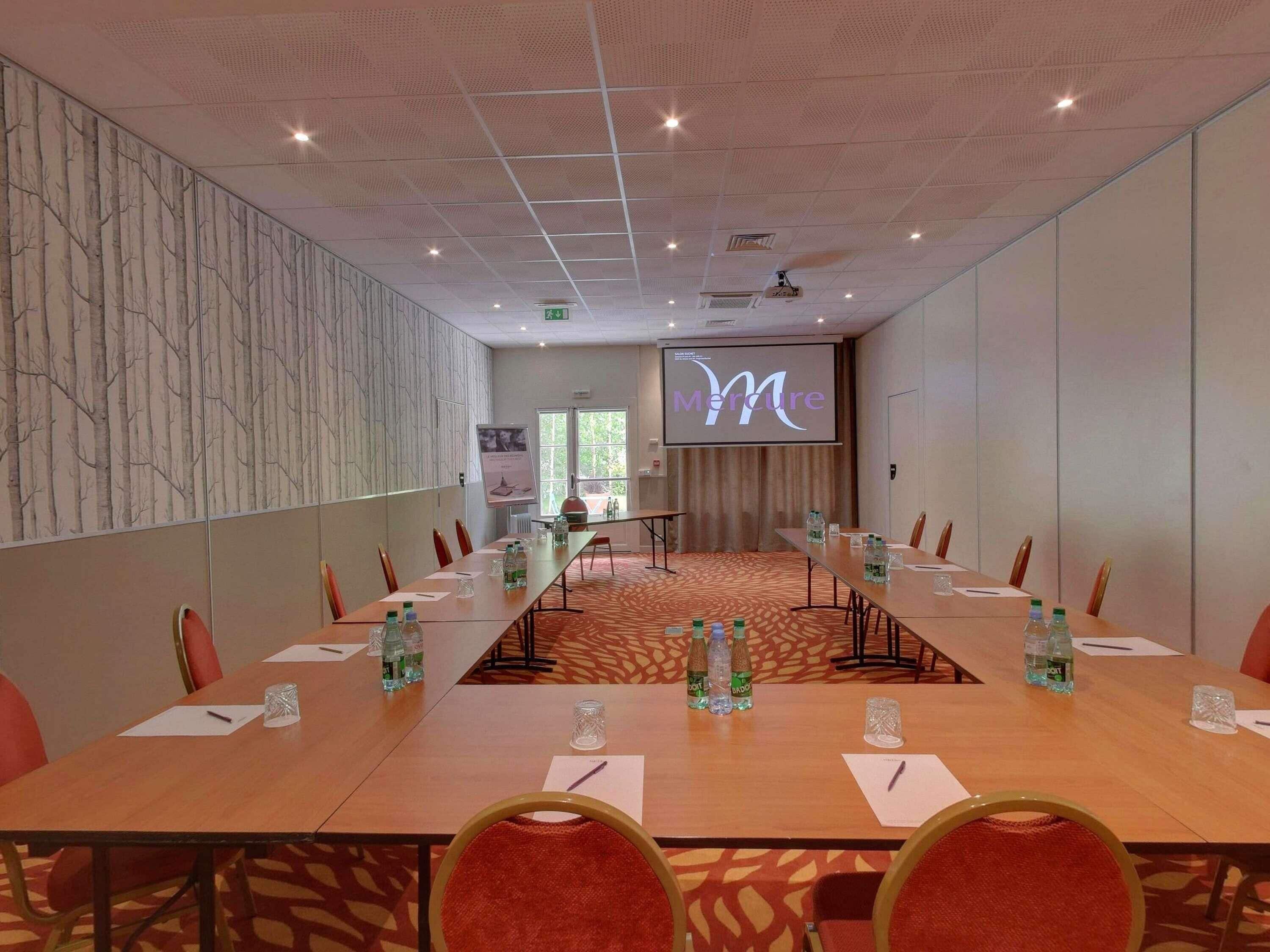 A conference room with a long table, chairs, water bottles, and a screen displaying the word 'Mercure'.
