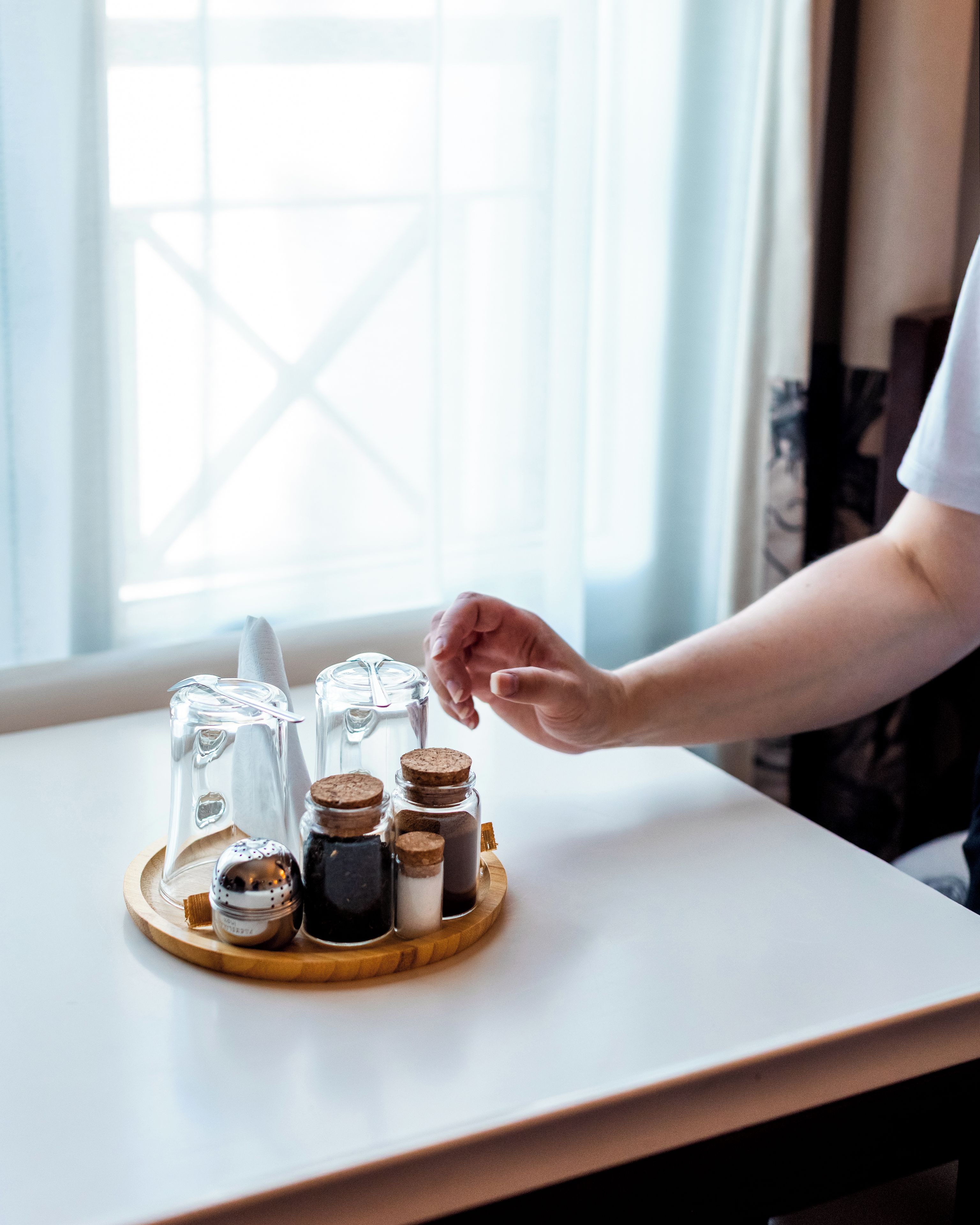 Una mesa con una bandeja que sostiene varios recipientes, entre ellos un salero, un pimentero y un frasco pequeño, en una habitación con una ventana al fondo.