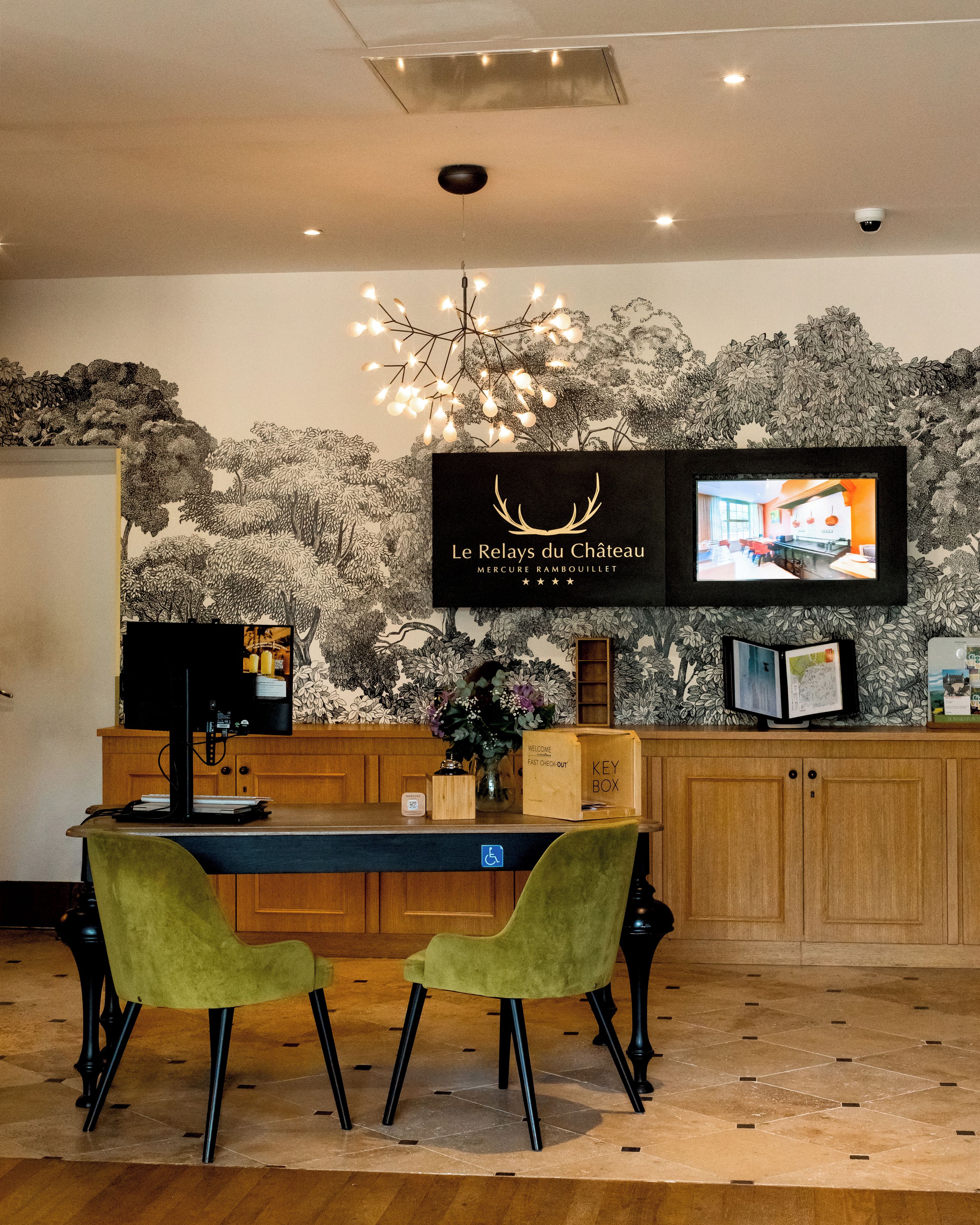 Un espace d’accueil avec un bureau en bois, deux chaises vertes, un téléviseur fixé au mur et un mur décoratif orné d’une illustration d’arbre.